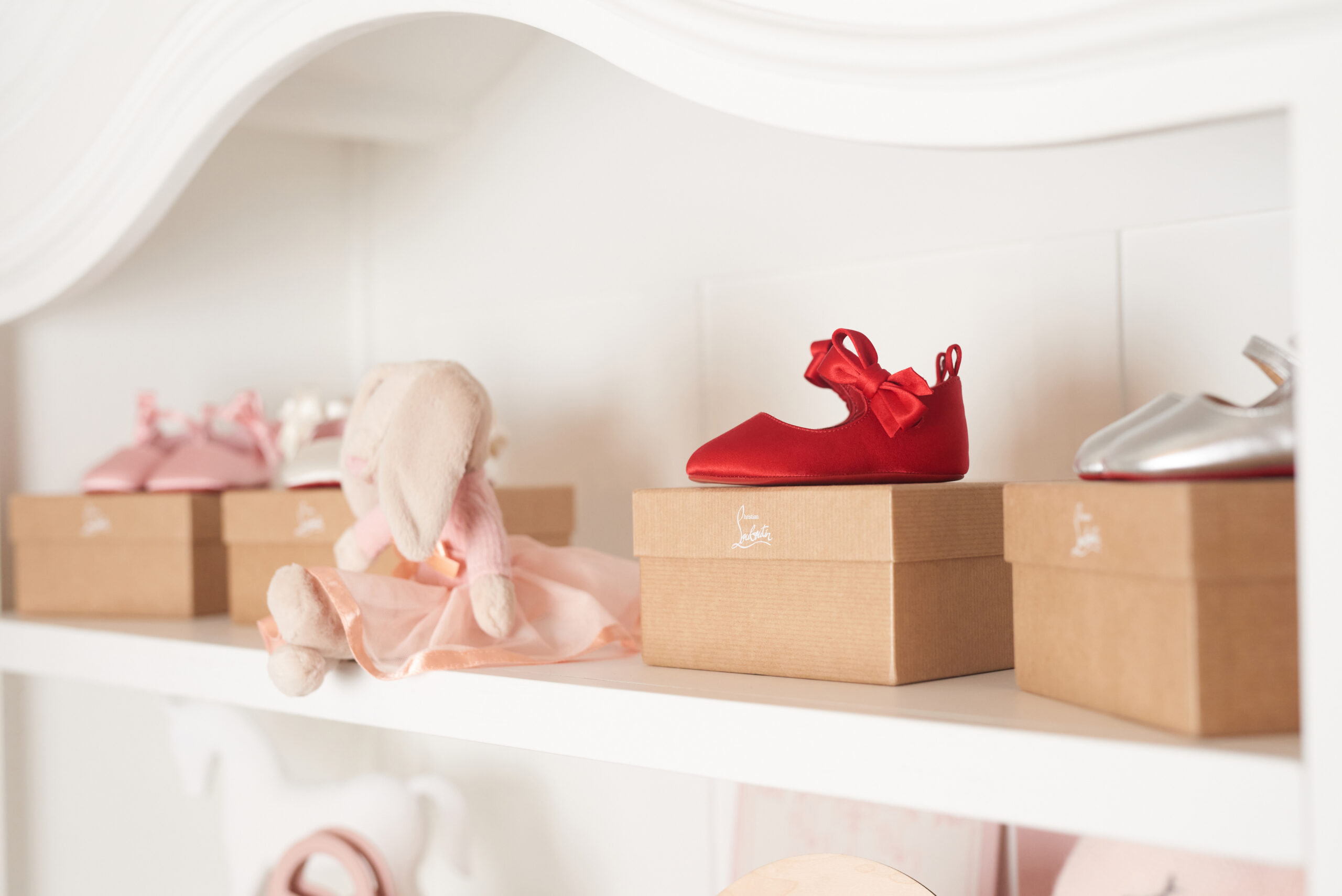 A tiny red bow shoe on a Christian Louboutin box beside a blush bunny, pink baby shoes, and a silver baby shoe.