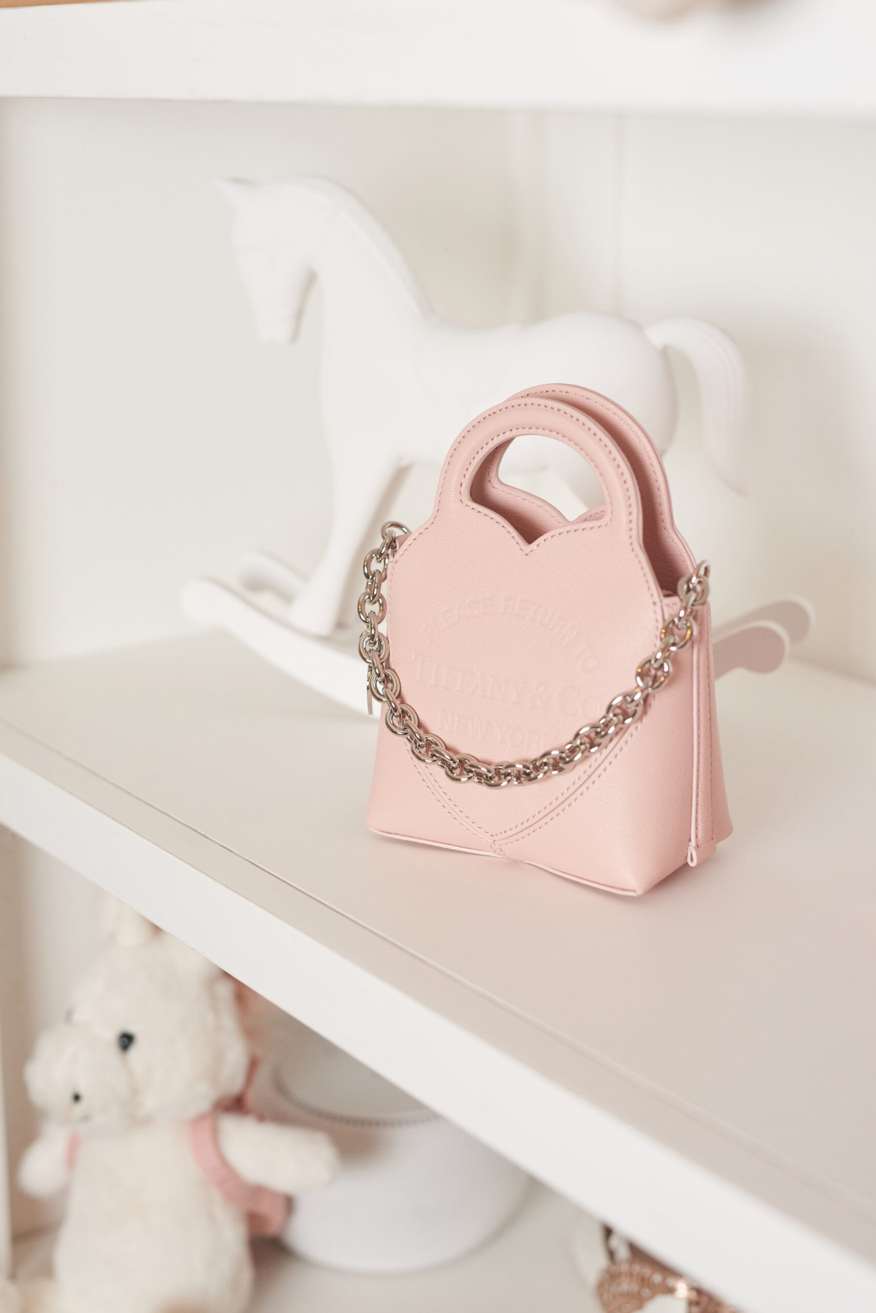A miniature blush-pink bag with a silver chain on a white shelf in front of a white rocking horse.