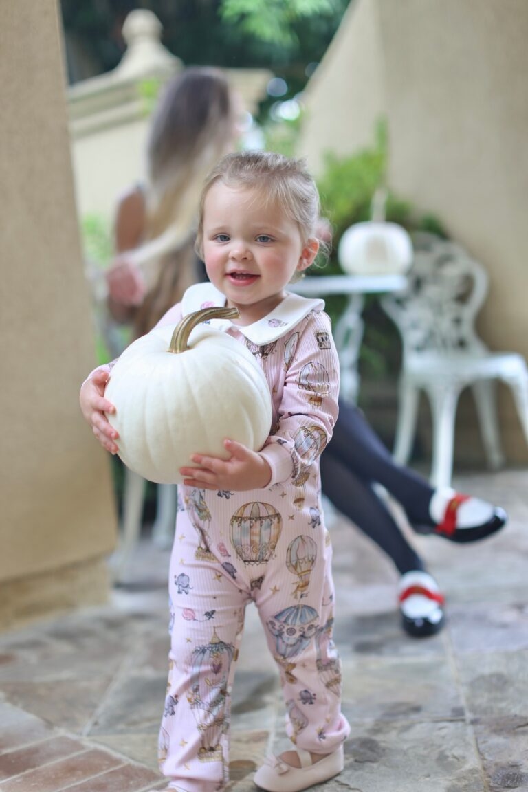 Madison in pink printed pajamas holding a white pumpkin on a patio.