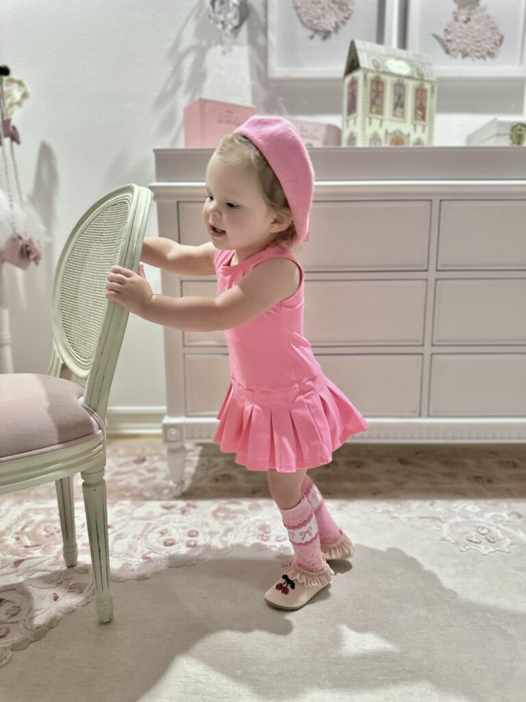 Madison wearing a pink dress, matching beret and pink socks beside a chair in her blush nursery.