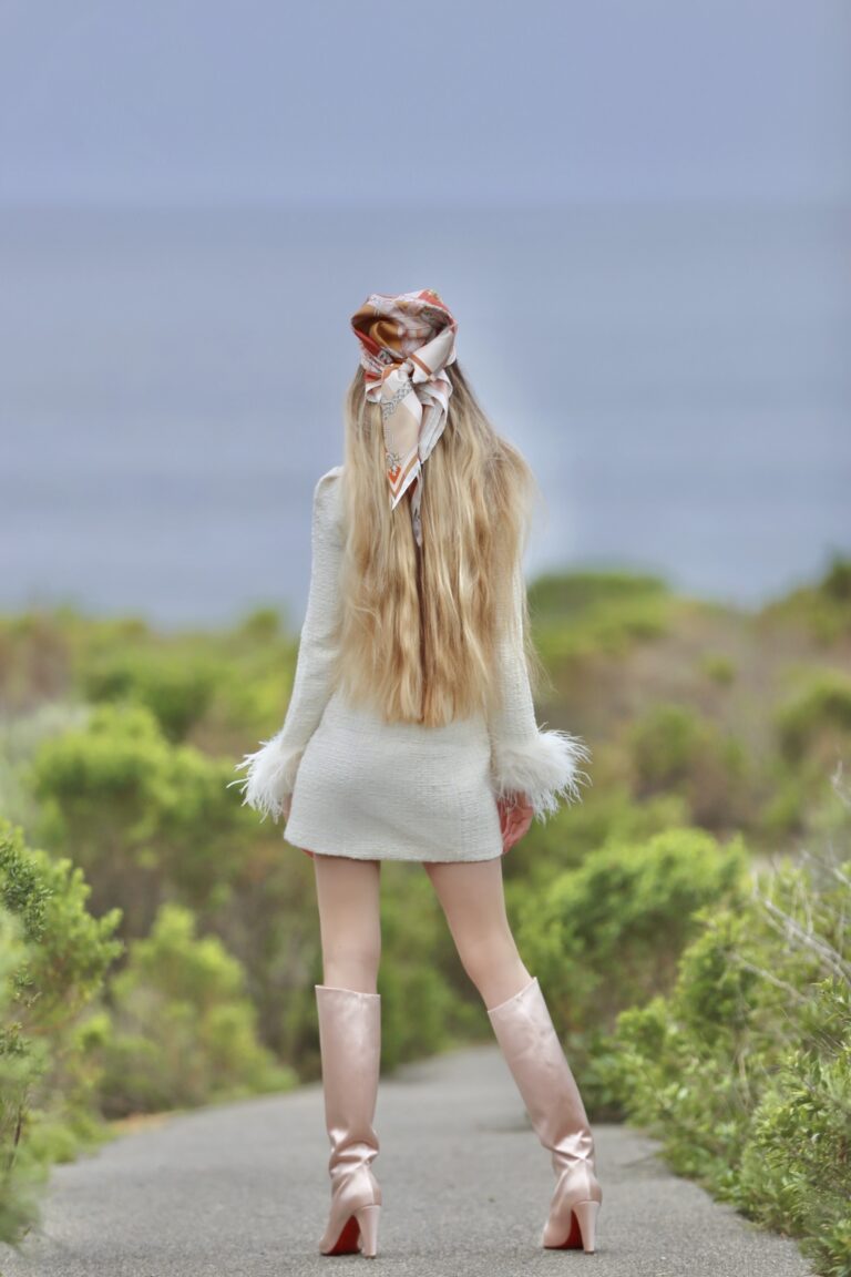 Madison from behind in an ivory dress, pink knee-high boots and a bow in her blonde hair on a garden path.