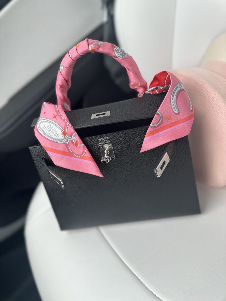 A black handbag with a pink silk scarf wrapped around its handle, resting on an ivory chair.