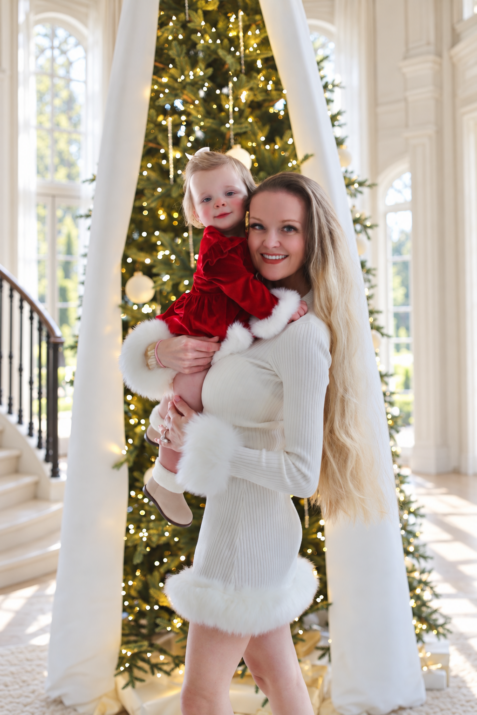Charlotte holding her daughter beside the Christmas tree, in ivory knit and red velvet.