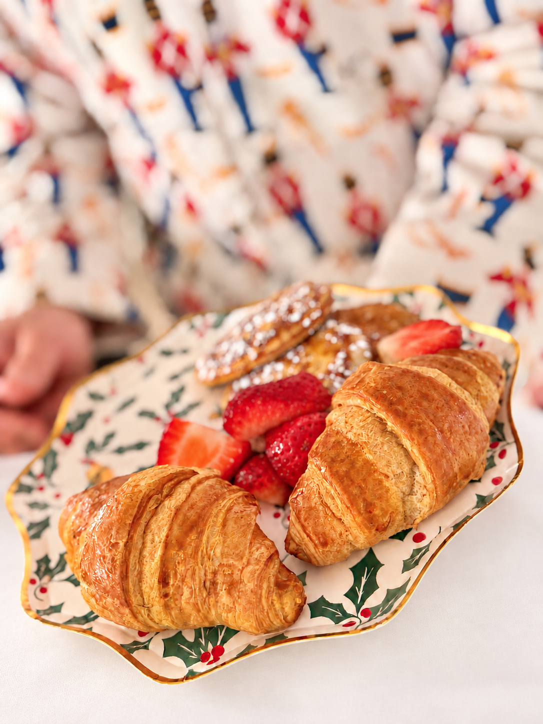 Croissants and strawberries on a holly-patterned Christmas platter above a soft white tablecloth, with gently blurred nutcracker pajamas.