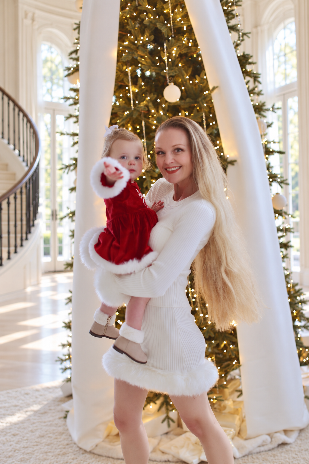 Charlotte holding her daughter, who reaches toward the camera beside the ribbon-dressed tree.