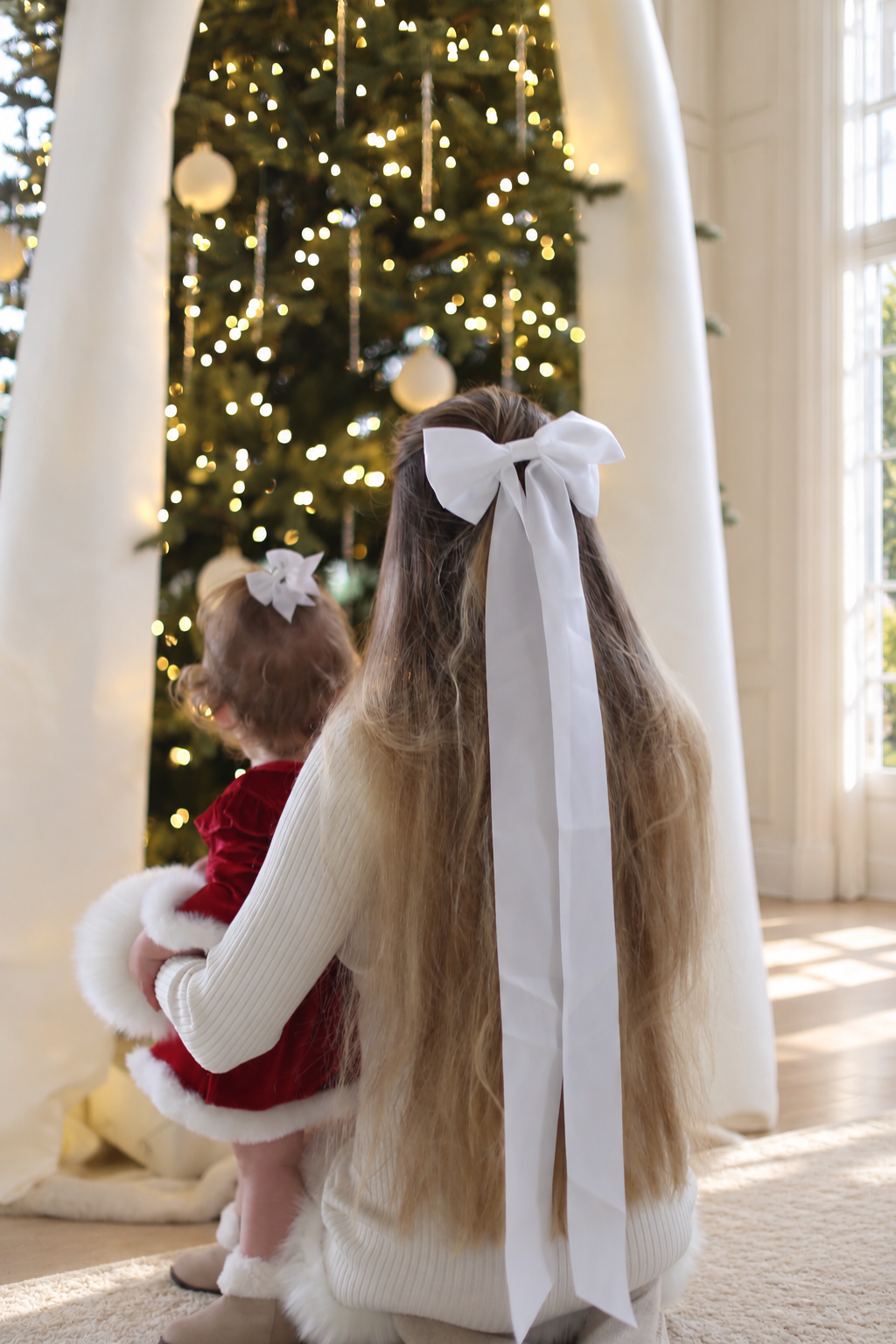 Rear view of Charlotte and her daughter wearing white hair bows beside the tree with its wide ivory ribbons.
