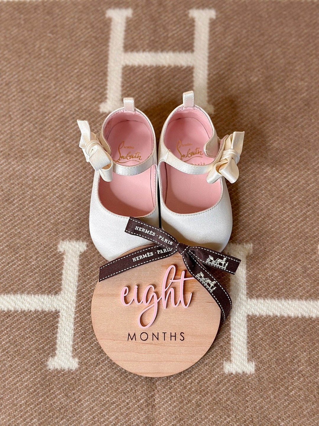 Ivory bow baby shoes beside an eight-month wooden milestone disc and brown Hermès ribbon, on an H-patterned blanket.