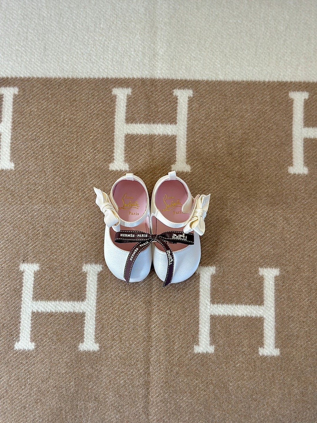 Ivory Christian Louboutin baby shoes with satin bows and a brown Hermès ribbon on a camel-and-ivory H-patterned blanket.