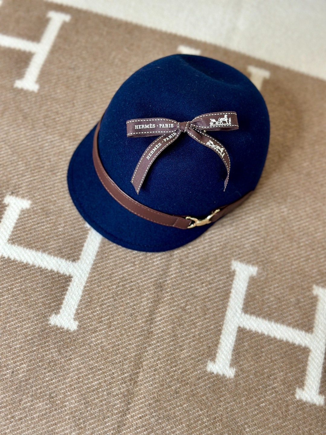A navy cap with a brown leather band, gold-tone hardware, and a brown Hermès ribbon bow on a camel-and-ivory H-patterned blanket.